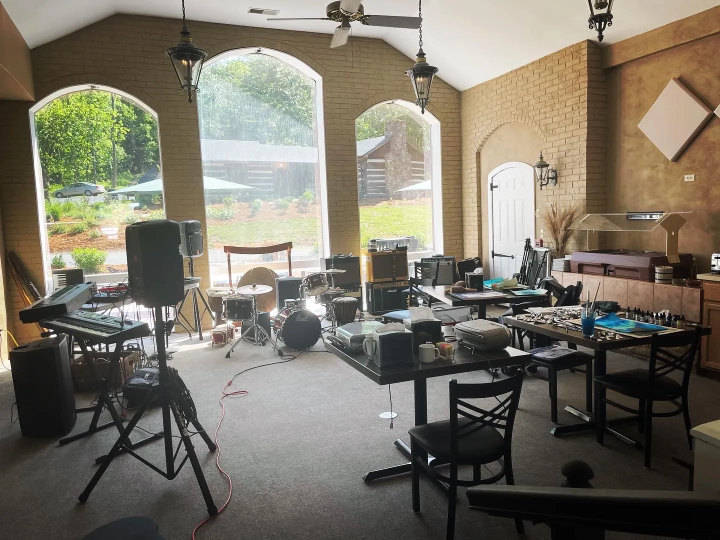 A large room set up with keyboards, speakers, a drum kit and tables of recording gear in front of arched windows