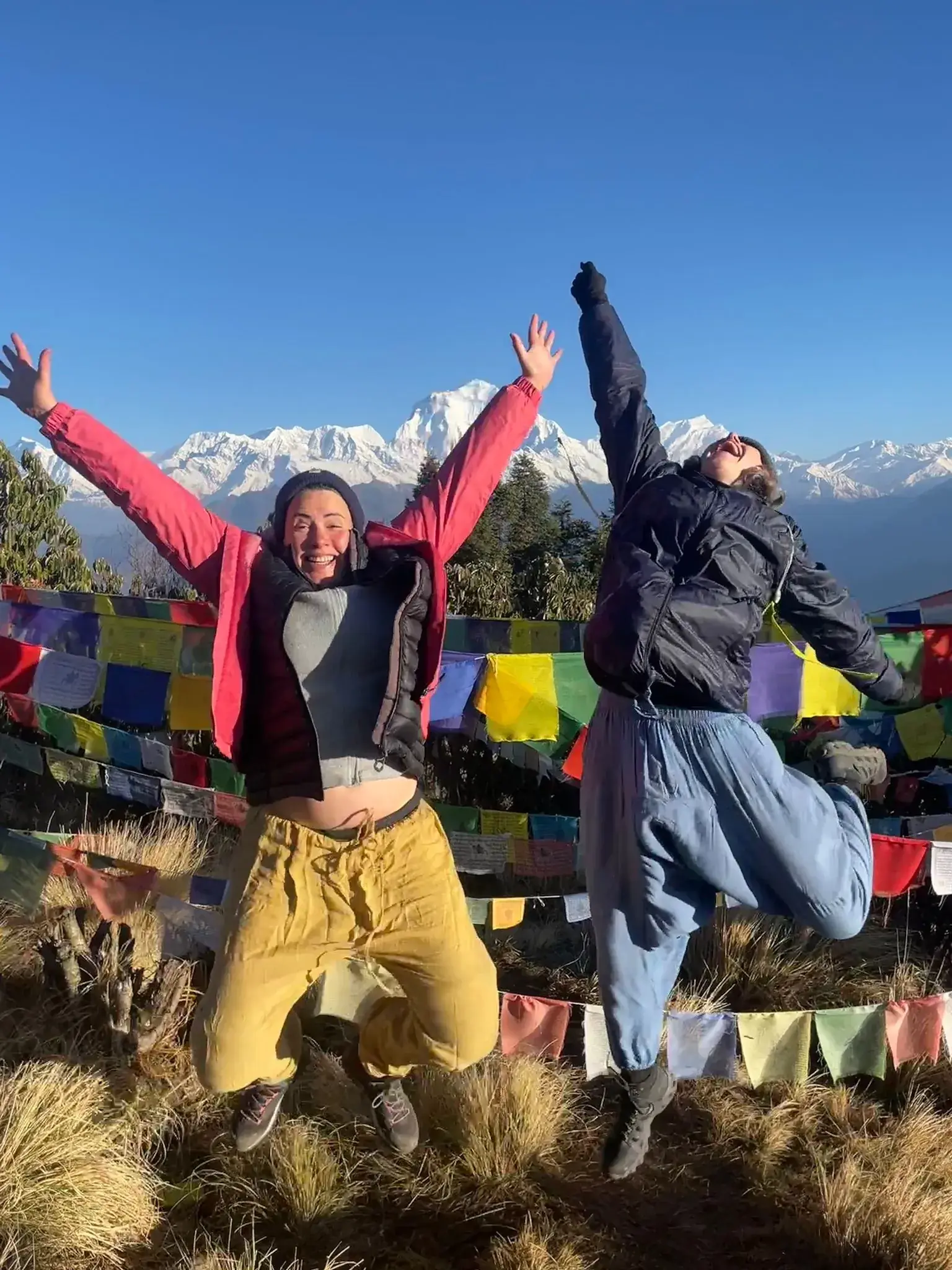 Caz and Laura jumping in the air in front of prayer flags and snow-capped Himalayan peaks